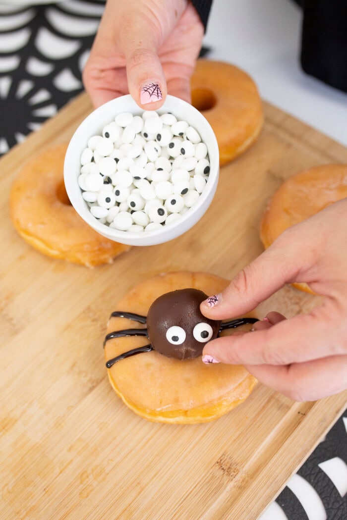 A person decorates a donut with a chocolate spider and candy eyes while holding a bowl of candy eyes in the other hand. More plain donuts are arranged on a wooden board, showcasing these festive Halloween donuts.