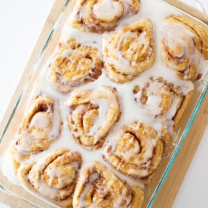 A glass baking dish filled with freshly baked cinnamon rolls covered in creamy white icing. The rolls are arranged in rows, nestled together on a wooden cutting board. The glaze appears glossy and is slightly melting into the rolls.