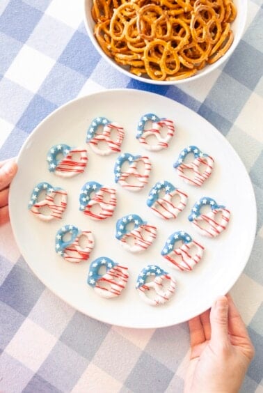 A plate of pretzels decorated with red, white, and blue icing to resemble the American flag, held above a blue-and-white checkered tablecloth. A bowl of plain pretzels sits nearby.