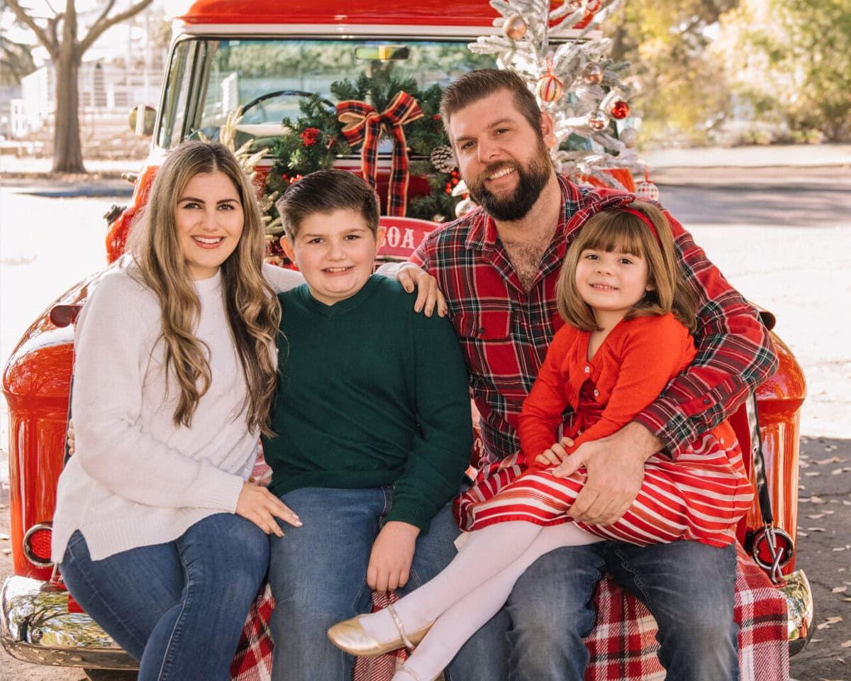 A smiling family of four sits on the tailgate of a red vintage truck decorated with Christmas ornaments and wreaths. The parents, son, and daughter are dressed in festive, casual clothes. Trees and sunlight are in the background.