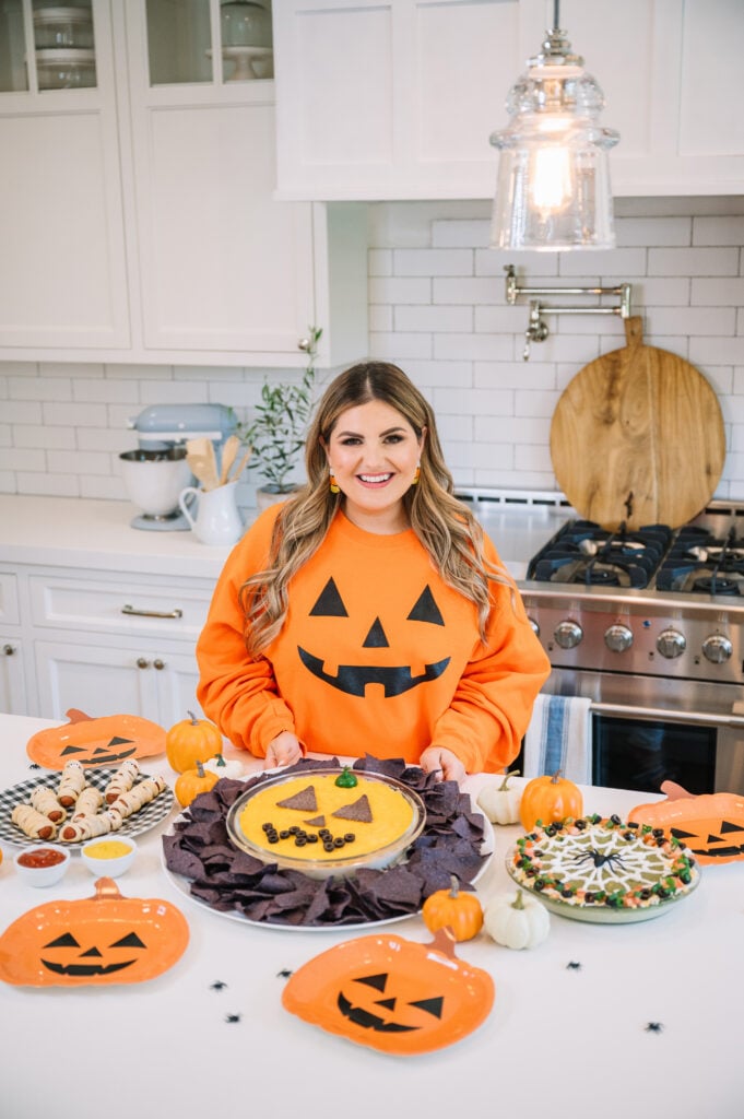 A woman in a pumpkin-themed sweatshirt stands in a bright kitchen, smiling behind a table with Halloween-themed dishes, including a dip with a jack-o-lantern face and pumpkin-shaped plates.