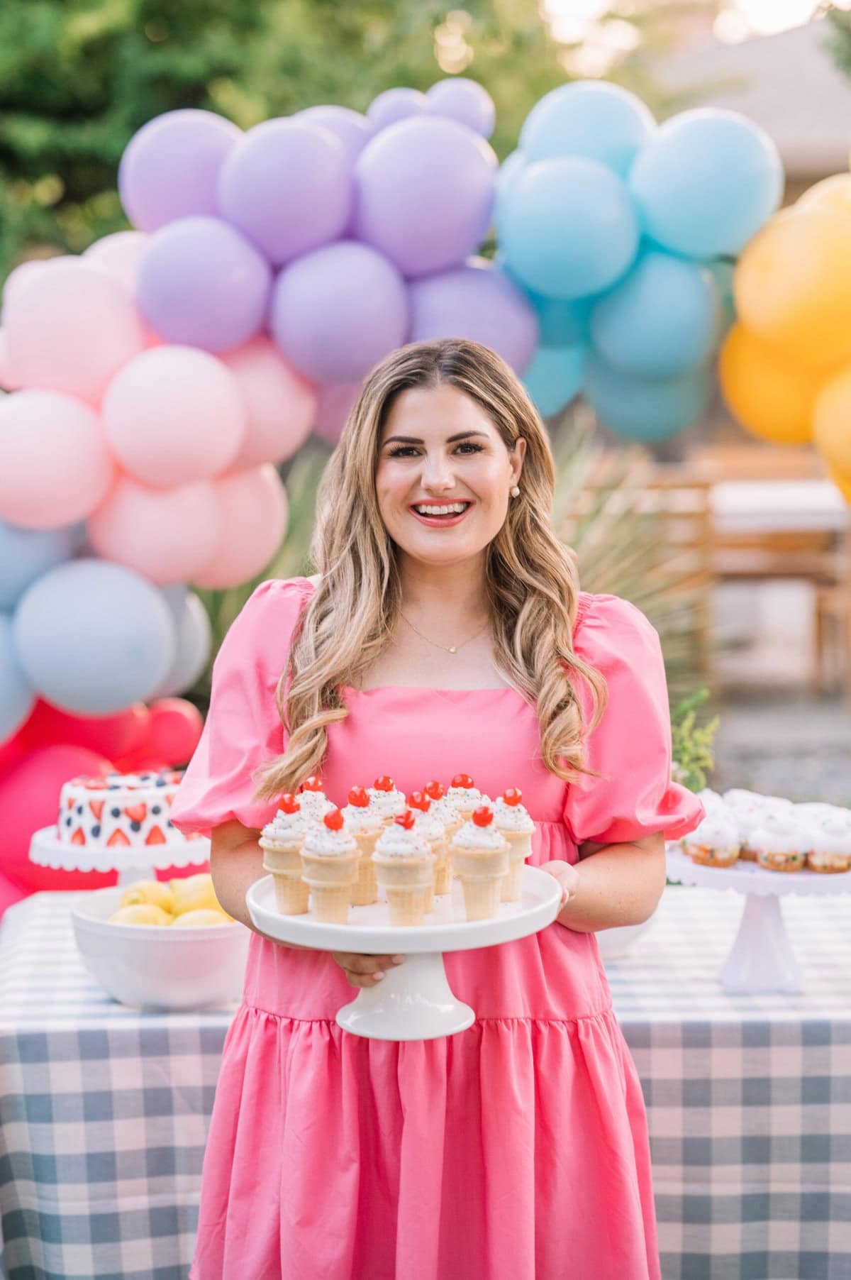 A woman in a bright pink dress smiles while holding a tray of cupcakes topped with cherries. She stands in front of a table with desserts and colorful balloon decorations outdoors.