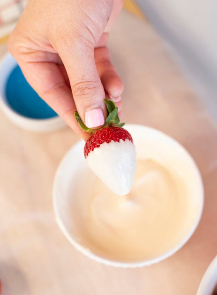 A hand with pink nail polish holds a strawberry partially dipped in white chocolate above a white bowl filled with melted white chocolate.