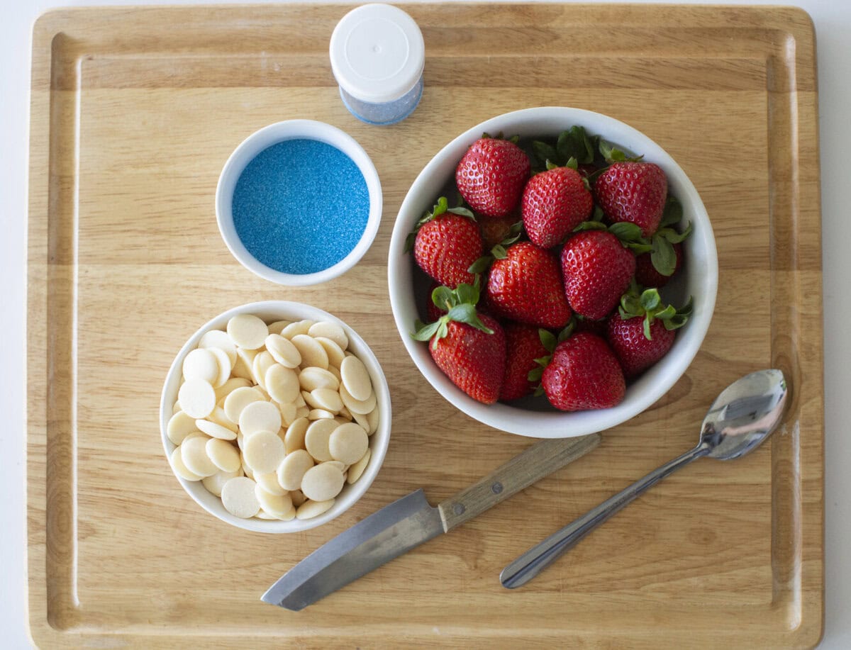 A wooden cutting board with a bowl of fresh strawberries, a bowl of white chocolate melts, a bowl of blue sprinkles, a container of blue sprinkles, a metal spoon, and a knife.