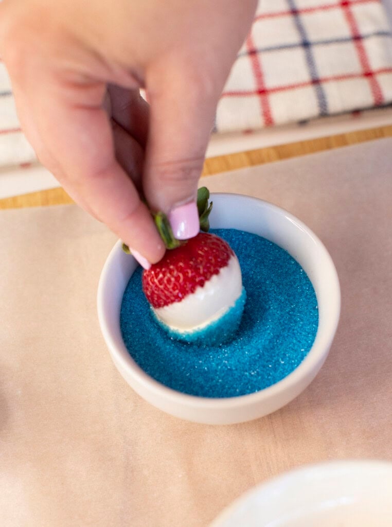 A hand dips a white chocolate-covered strawberry into a bowl of blue sugar, creating a red, white, and blue treat. A striped cloth is visible in the background.