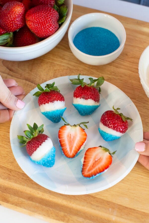 A plate of strawberries dipped in white chocolate and blue sugar sits on a wooden surface. Two hands hold the plate. A bowl of plain strawberries, a bowl of blue sugar, and a bowl of white chocolate are nearby.