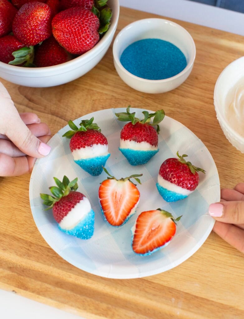 A plate of strawberries dipped in white chocolate and blue sugar sits on a wooden surface. Two hands hold the plate. A bowl of plain strawberries, a bowl of blue sugar, and a bowl of white chocolate are nearby.