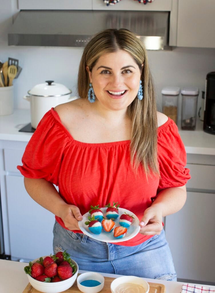 A woman in a red off-the-shoulder top holds a plate of decorated strawberries in a modern kitchen. Bowls of strawberries, blue sprinkles, and white chocolate are on the counter in front of her.