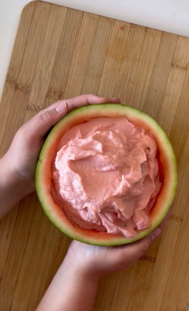 A pair of hands holding a hollowed-out watermelon half filled with pink whipped mixture, resting on a wooden cutting board—an easy summer dessert perfect for your list of summer dessert ideas.