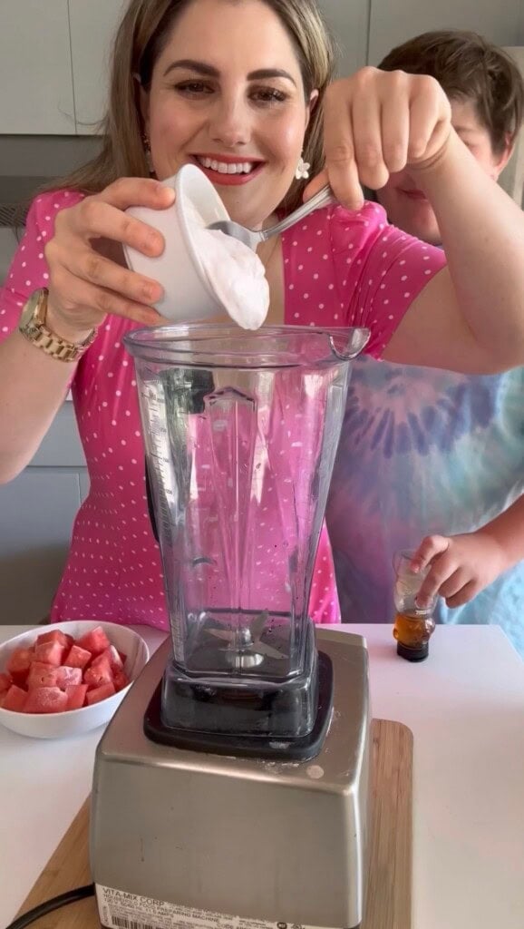 A woman in a pink polka dot dress smiles while spooning yogurt into a blender, making easy summer desserts. A child next to her holds a small glass with liquid, and a bowl of watermelon pieces sits on the counter.