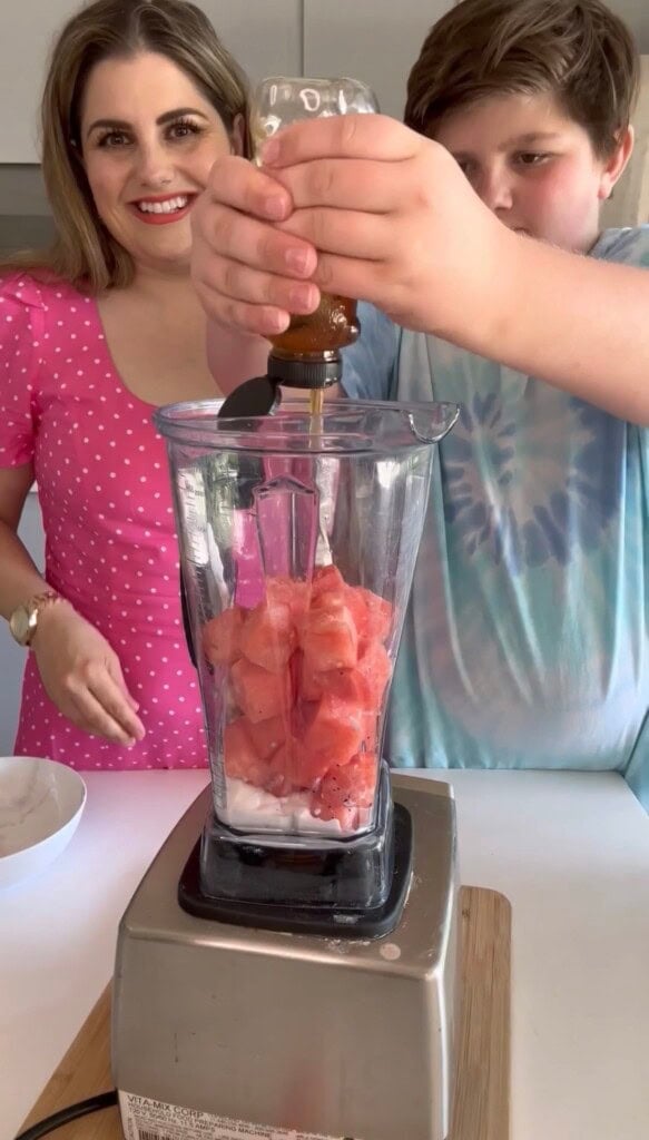 A woman in a pink polka dot dress and a boy in a tie-dye shirt stand by a blender filled with watermelon cubes, making an easy summer dessert as the boy pours honey in and the woman smiles.