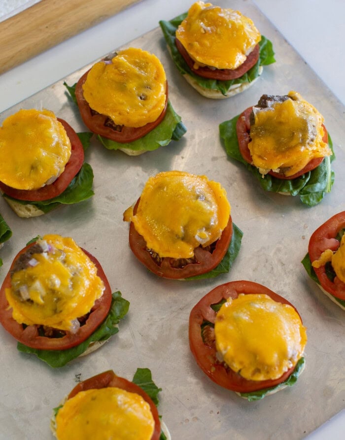 Several open-faced mini burgers are arranged on a baking sheet, each topped with a lettuce leaf, tomato slice, cooked beef patty, and melted cheddar cheese.