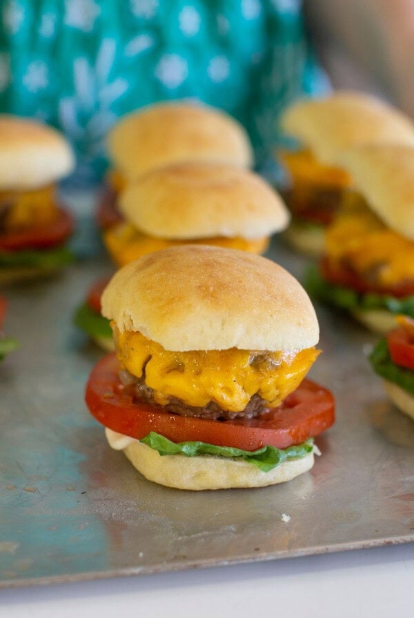 Mini cheeseburgers with lettuce, tomato, and melted cheddar on small buns are arranged on a metal tray. A person wearing a green patterned shirt is visible in the background.
