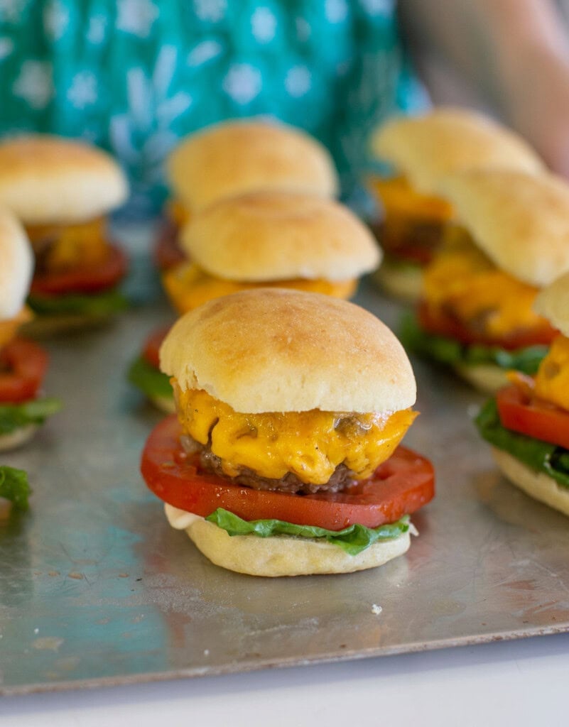 Mini cheeseburgers with lettuce, tomato, and melted cheddar on small buns are arranged on a metal tray. A person wearing a green patterned shirt is visible in the background.