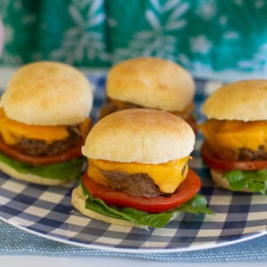 Four cheeseburger sliders with lettuce and tomato on buns are arranged on a blue-and-white checkered plate. A person in a green patterned shirt is visible in the background.