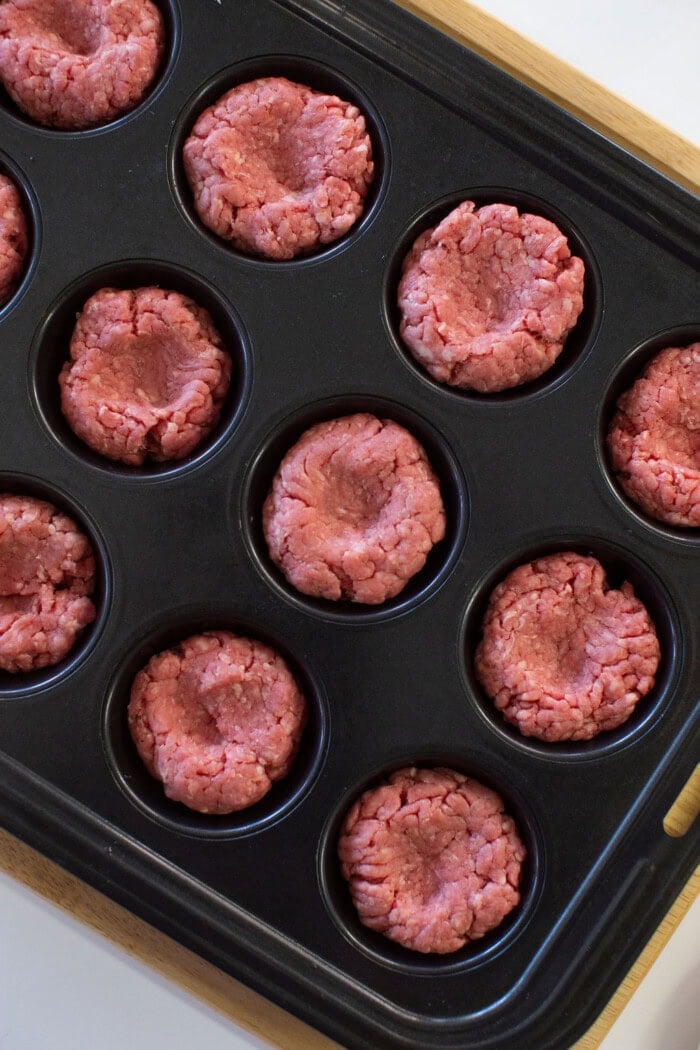 A muffin tin filled with raw ground beef pressed into cup shapes, ready to be cooked.
