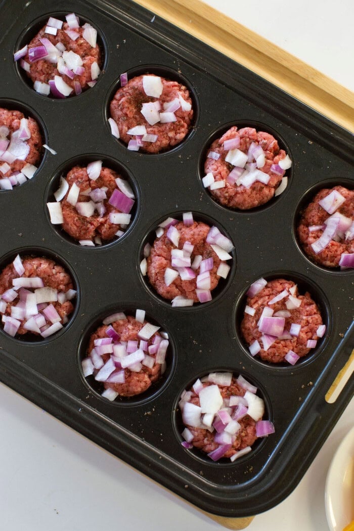 A muffin tin filled with raw ground beef topped with chopped red onions, ready to be cooked, is placed on a wooden board.