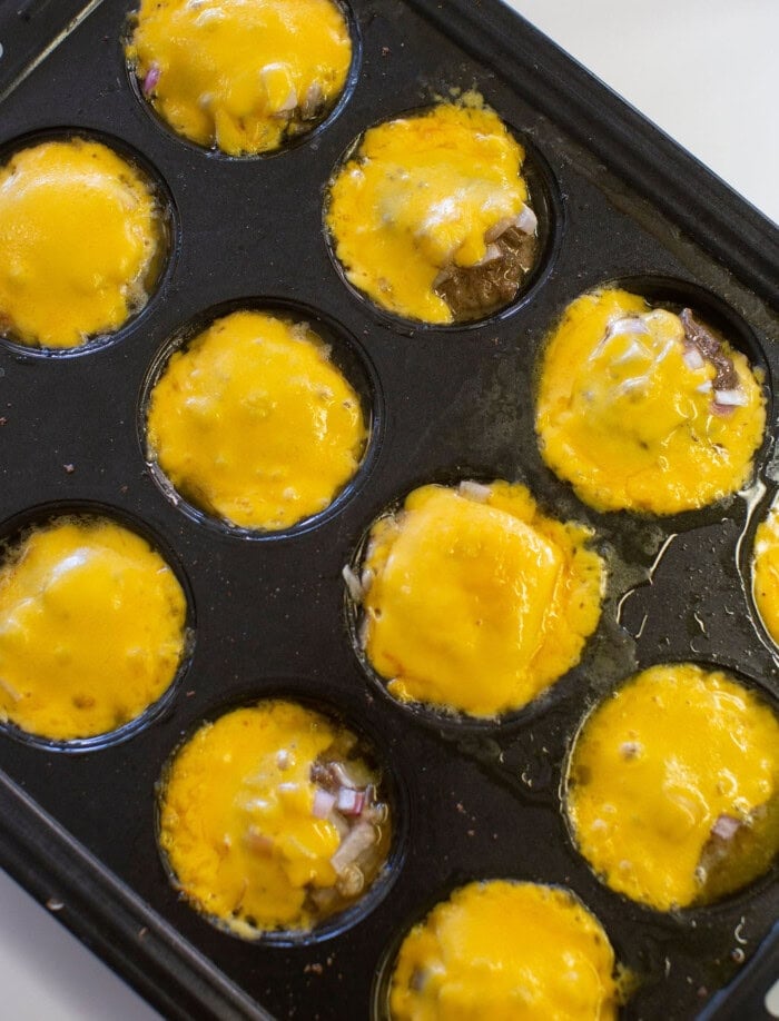 Muffin tin filled with baked mini meat patties topped with melted yellow cheese, some with visible chopped onions, resting on a white surface.