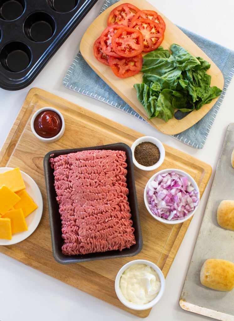 A wooden board holds ground beef, sliced red onions, mayonnaise, and cheese slices. Nearby are sliced tomatoes and spinach, a muffin tin, ketchup, pepper, and partially visible sandwich buns on a baking sheet.