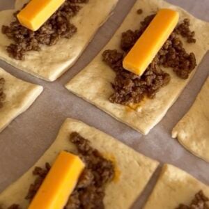 Rectangular pieces of dough topped with cooked ground beef and a stick of cheddar cheese, arranged on parchment paper and ready to be wrapped or baked—perfect for easy dinner recipes for family.