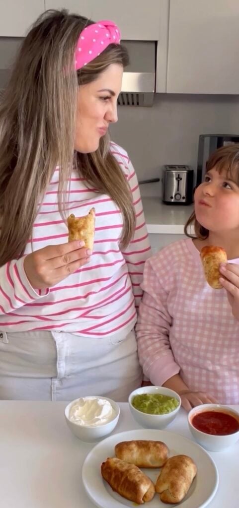 A woman and a young girl wearing pink and white tops smile at each other while holding pastries in a kitchen, surrounded by three dips and more pastries—perfect for easy dinner ideas for family.