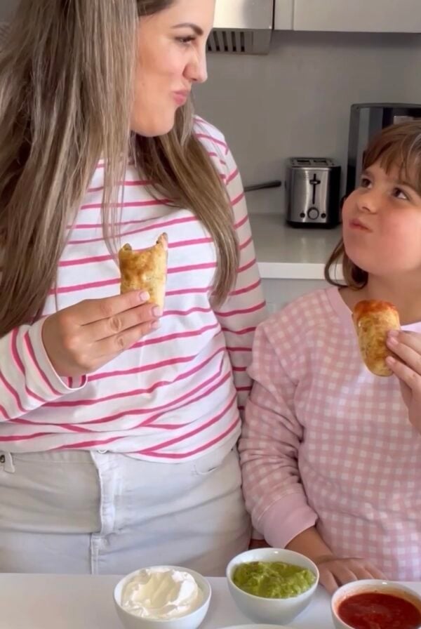 A woman and a young girl wearing pink and white tops smile at each other while holding pastries in a kitchen, surrounded by three dips and more pastries—perfect for easy dinner ideas for family.