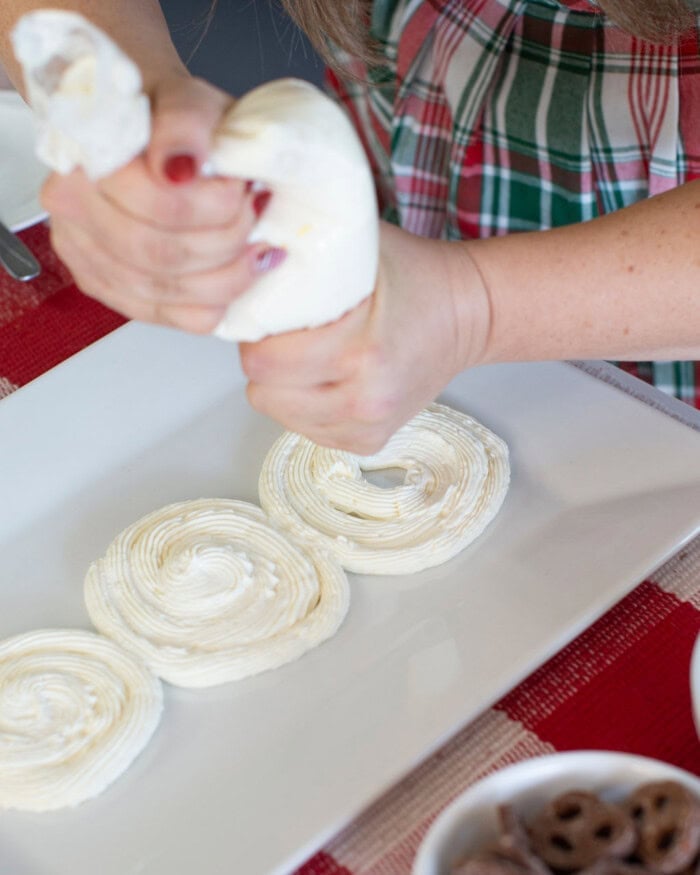 A person wearing a plaid shirt pipes swirls of white icing onto a rectangular plate covered with a red and white placemat. A small bowl with chocolate pretzels is partially visible in the corner.