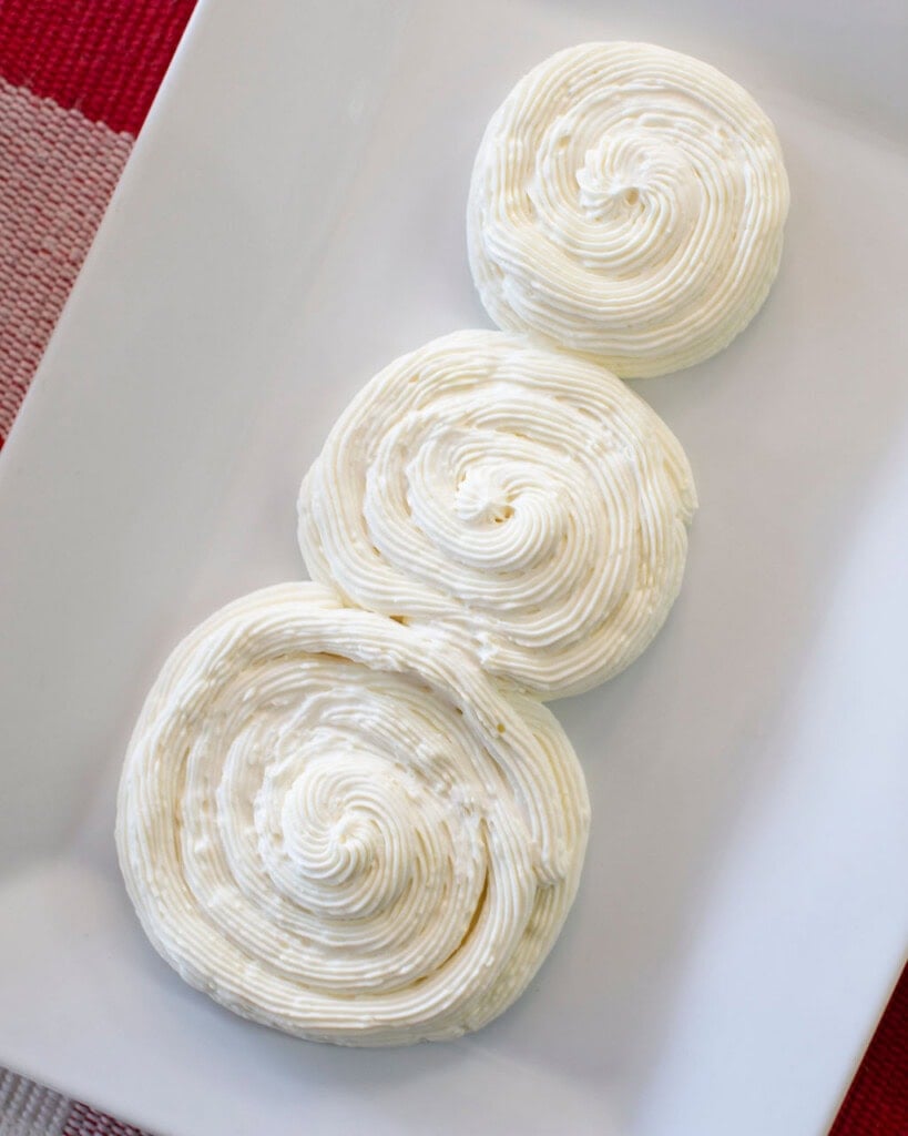 Three swirls of piped white frosting arranged vertically on a white rectangular plate, resembling a snowman shape. The background includes part of a red and white checkered cloth.
