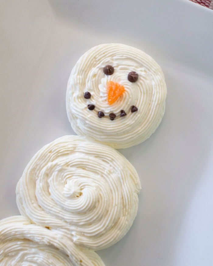 A snowman made of piped white frosting on a white surface, with chocolate chips for eyes and mouth, and an orange frosting nose.