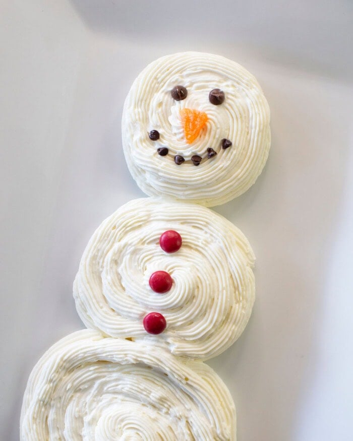 A snowman made of three round, white frosted desserts is decorated with chocolate chips for eyes and mouth, an orange candy for a nose, and three red candies as buttons, arranged on a white plate.
