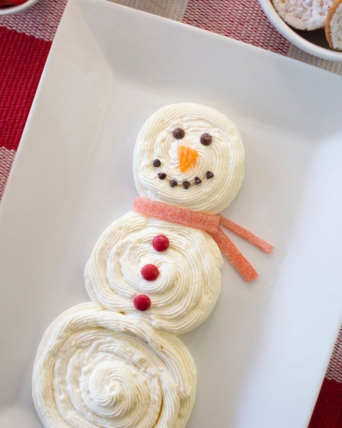 A snowman-shaped dessert made of swirled white frosting, decorated with chocolate chips for eyes and mouth, an orange candy nose, red candies as buttons, and a red gummy candy scarf, arranged on a white platter.