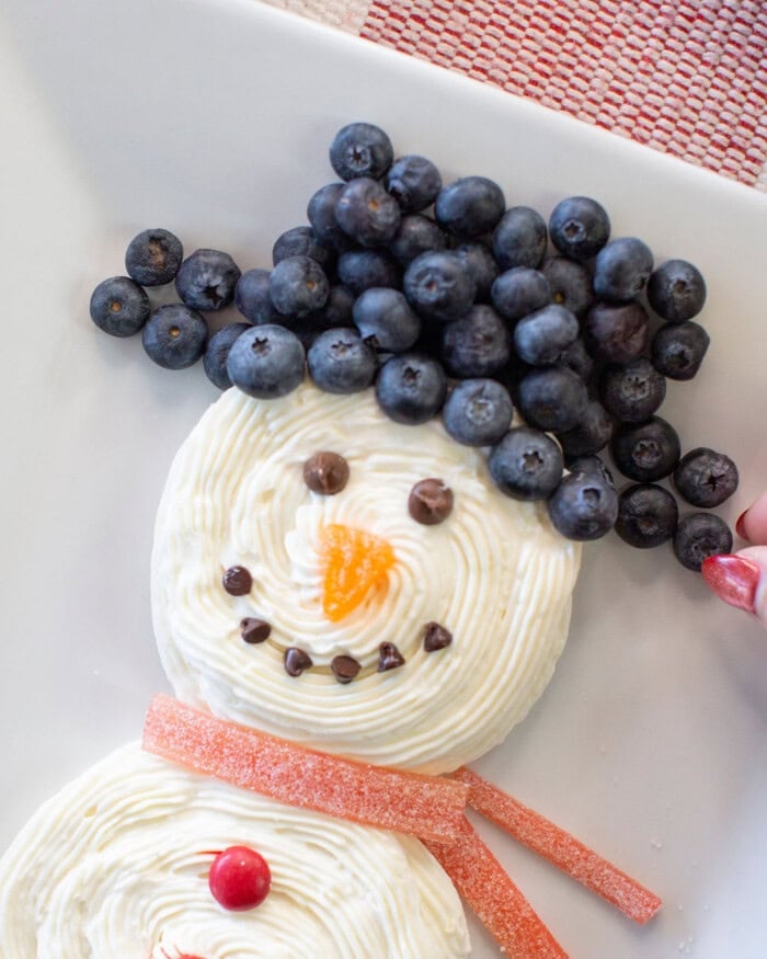 A dessert shaped like a snowman, decorated with blueberries for hair, frosting for the face, chocolate chips for the eyes and mouth, an orange candy nose, and red gummy strips as a scarf, on a white plate.
