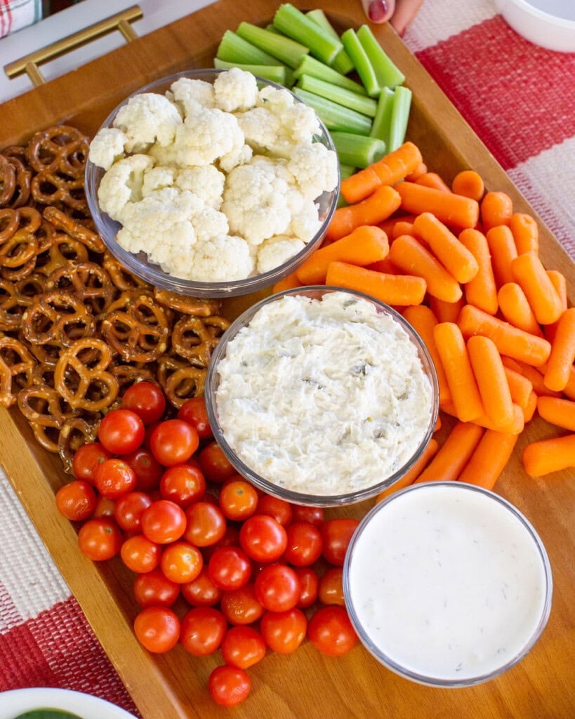 A wooden tray with bowls of dip, surrounded by cherry tomatoes, pretzels, celery sticks, cauliflower florets, and baby carrots, set on a red and white checkered tablecloth.