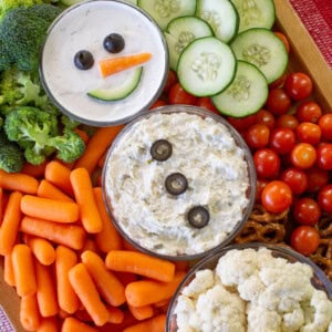 A festive veggie platter includes cherry tomatoes, carrots, broccoli, cauliflower, cucumbers, and two dips decorated as snowmen with olives for eyes and buttons and a carrot for a nose, surrounded by vegetables and pretzels.