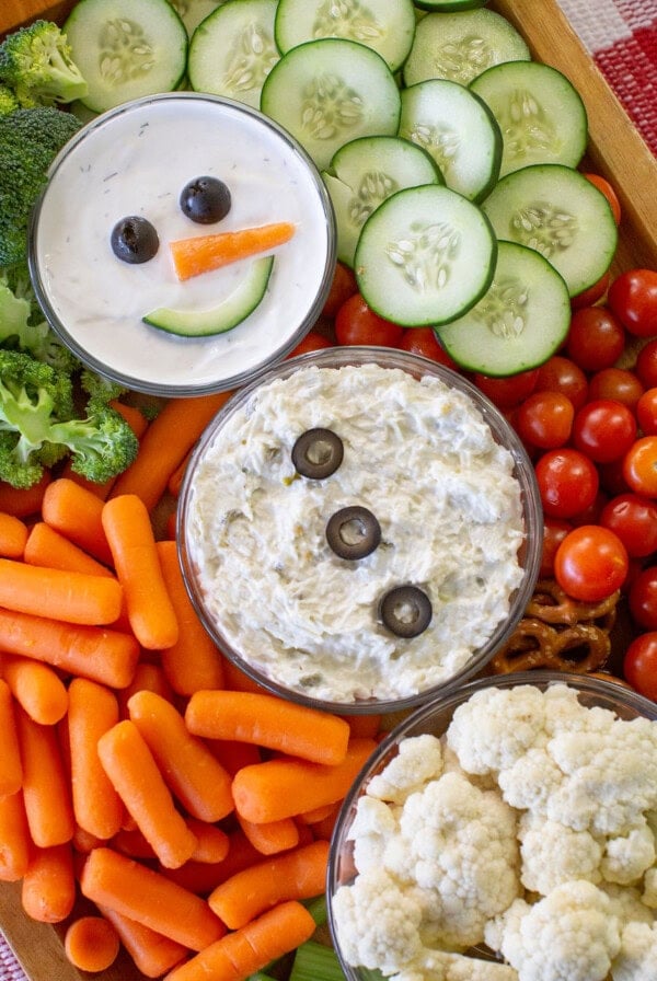 A festive veggie platter includes cherry tomatoes, carrots, broccoli, cauliflower, cucumbers, and two dips decorated as snowmen with olives for eyes and buttons and a carrot for a nose, surrounded by vegetables and pretzels.