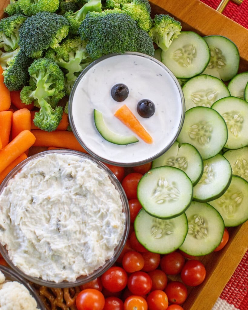 A platter of fresh vegetables surrounds a bowl of dip decorated as a snowman face with olive eyes, a carrot nose, and a cucumber slice smile. Sliced cucumbers, broccoli, carrots, and cherry tomatoes are visible.