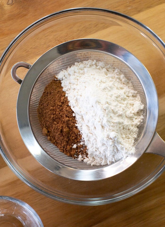 A metal sifter holds unsifted cocoa powder and flour over a glass mixing bowl on a wooden surface, ready for baking.