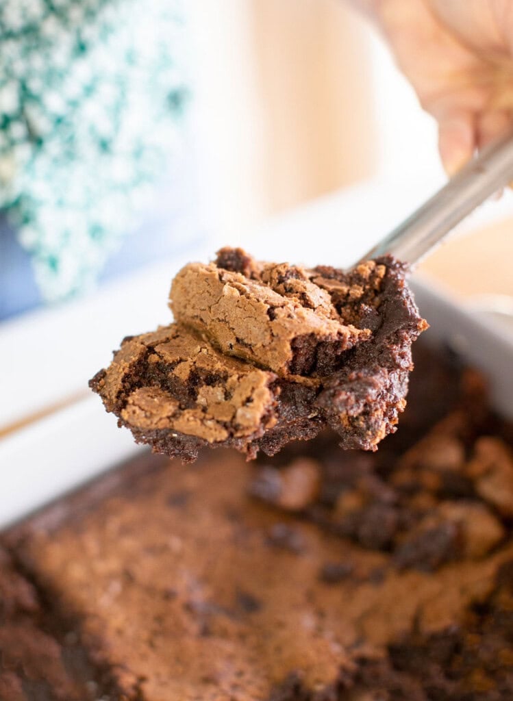 A hand holding a metal spatula lifts a gooey piece of chocolate brownie with a cracked top from a pan of brownies.