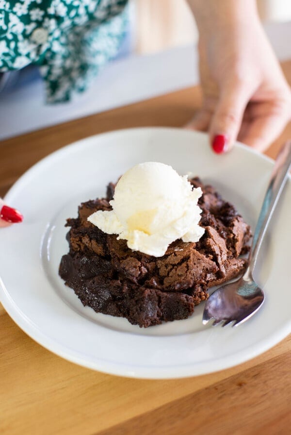 A person with red nail polish holds a white plate with a serving of chocolate brownie topped with a scoop of vanilla ice cream and a spoon beside it.