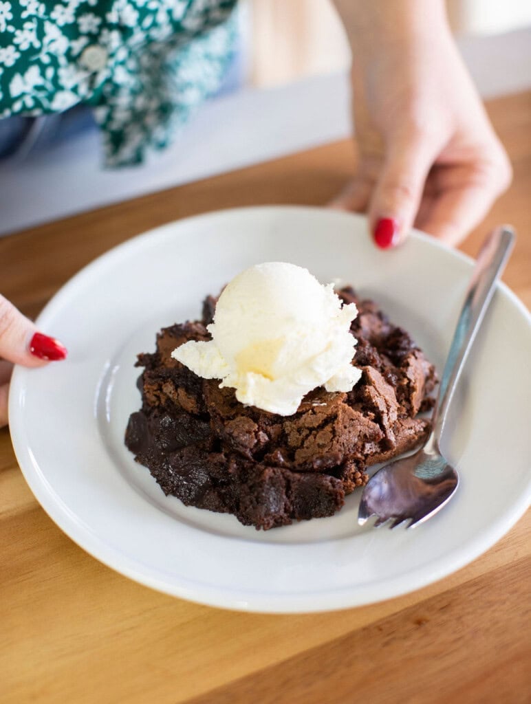 A person with red nail polish holds a white plate with a serving of chocolate brownie topped with a scoop of vanilla ice cream and a spoon beside it.