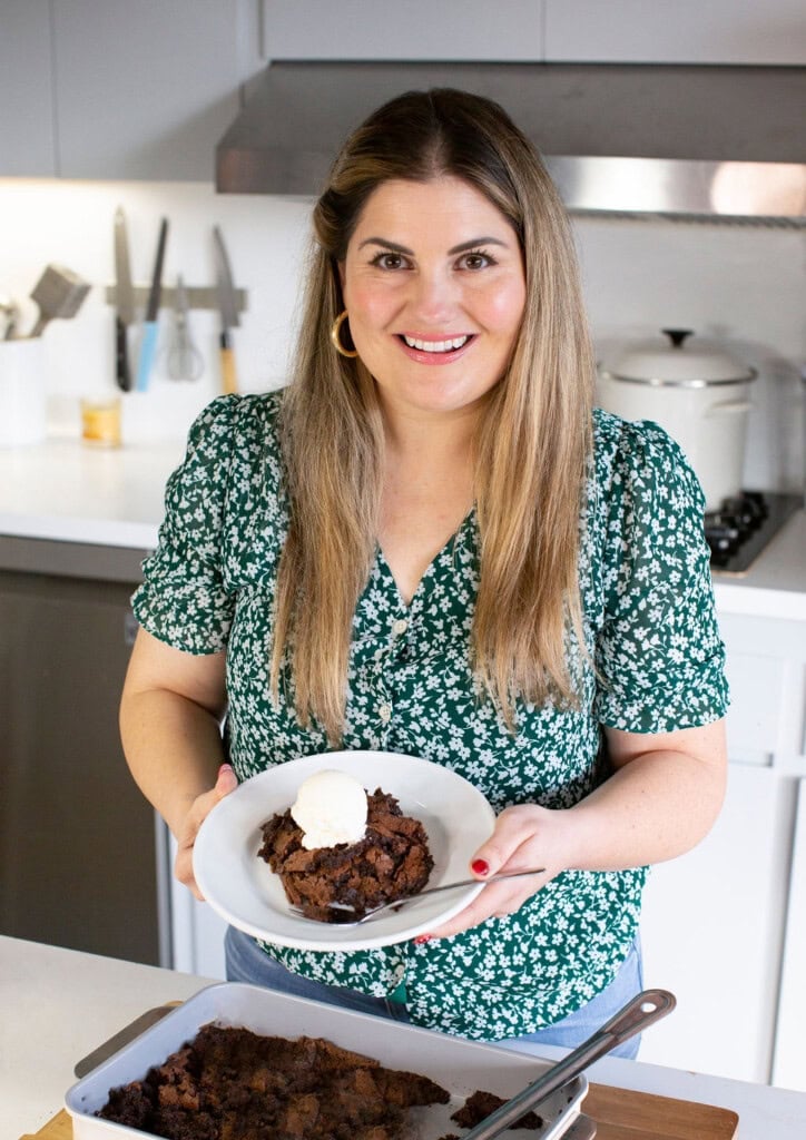 A woman with long hair, wearing a green floral blouse, stands in a kitchen holding a bowl of chocolate dessert topped with a scoop of vanilla ice cream. A pan of the same dessert is on the counter in front of her.