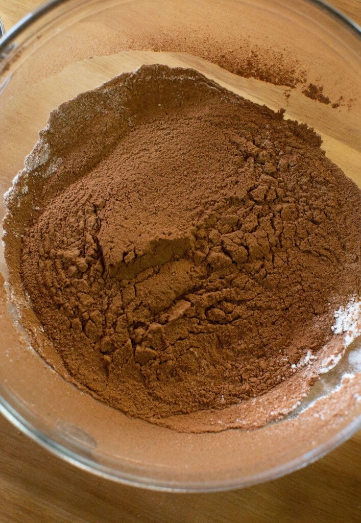 A glass mixing bowl filled with sifted cocoa powder and flour on a wooden surface, ready for baking.