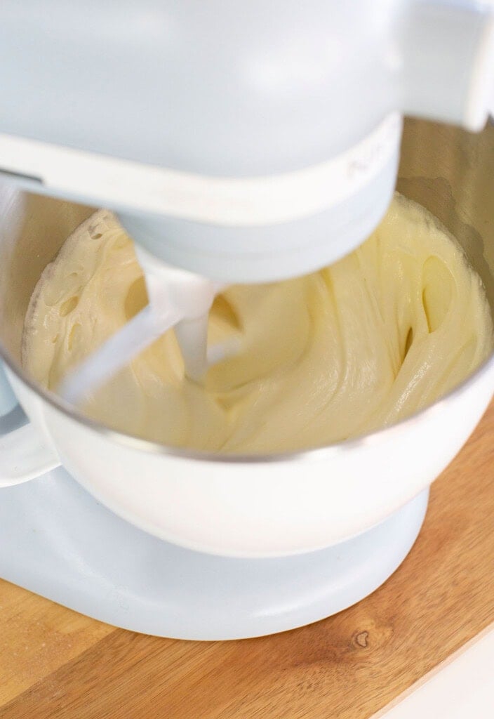 A white stand mixer blends a creamy batter in a metal mixing bowl, which is set on a light wooden countertop.
