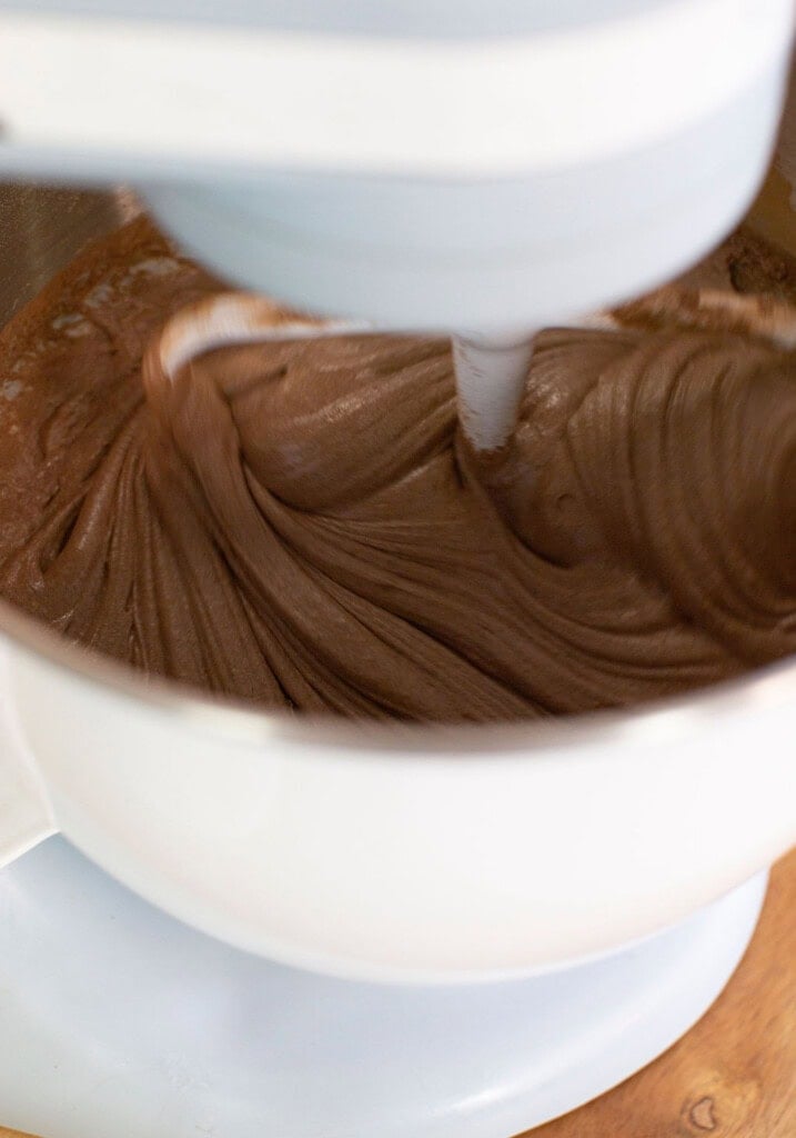 A close-up of a stand mixer blending chocolate batter in a white mixing bowl, with the mixer paddle in motion.
