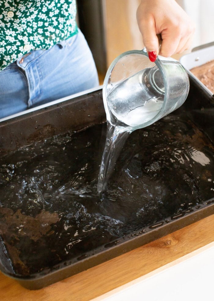 A person is pouring clear water from a glass pitcher into a large black baking pan on a wooden surface. The person is wearing jeans and a green floral top.