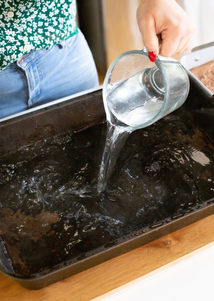 A person is pouring clear water from a glass pitcher into a large black baking pan on a wooden surface. The person is wearing jeans and a green floral top.