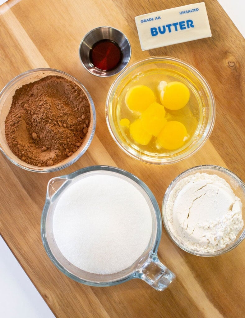 Five brownie ingredients arranged on a wooden surface: a bowl of cocoa powder, a bowl with three eggs, a stick of unsalted butter, a cup of sugar, a bowl of flour, and a small cup of vanilla extract.