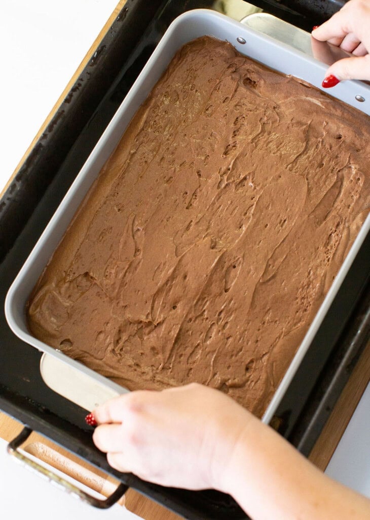 A person with red painted nails places a rectangular pan filled with chocolate cake batter into a larger black roasting pan.