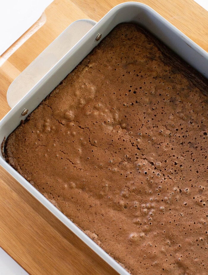A rectangular metal pan filled with freshly baked brownies sits on a wooden cutting board. The brownies have a cracked, slightly bubbly top and rich brown color.