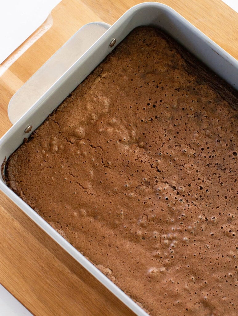 A rectangular metal pan filled with freshly baked brownies sits on a wooden cutting board. The brownies have a cracked, slightly bubbly top and rich brown color.